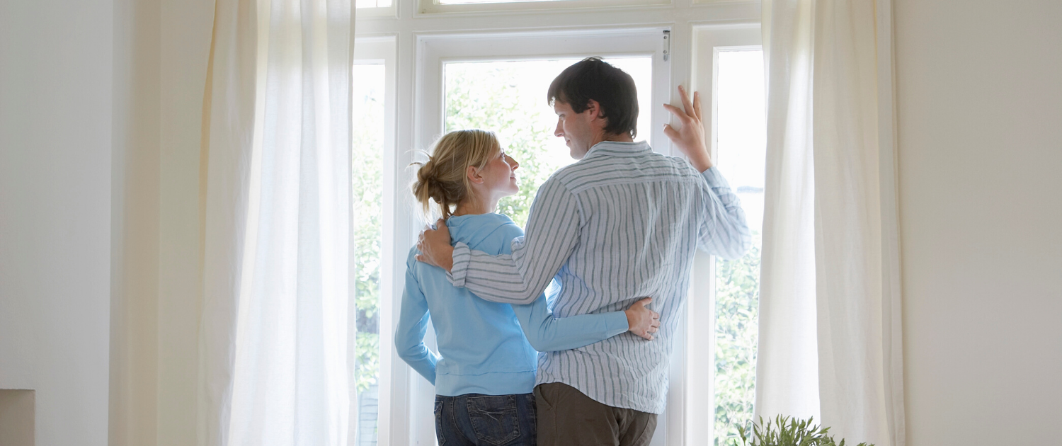 Two adults standing at home window, looking at one another with arms around each other